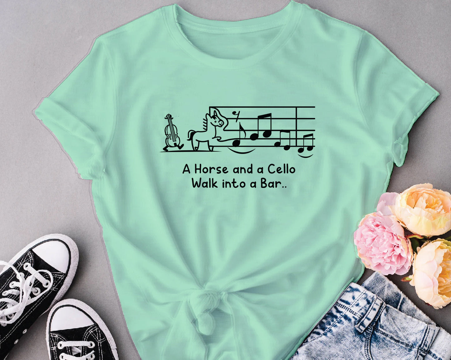 Mint green t-shirt with humorous design and text, placed on a gray surface with flowers and shoes.