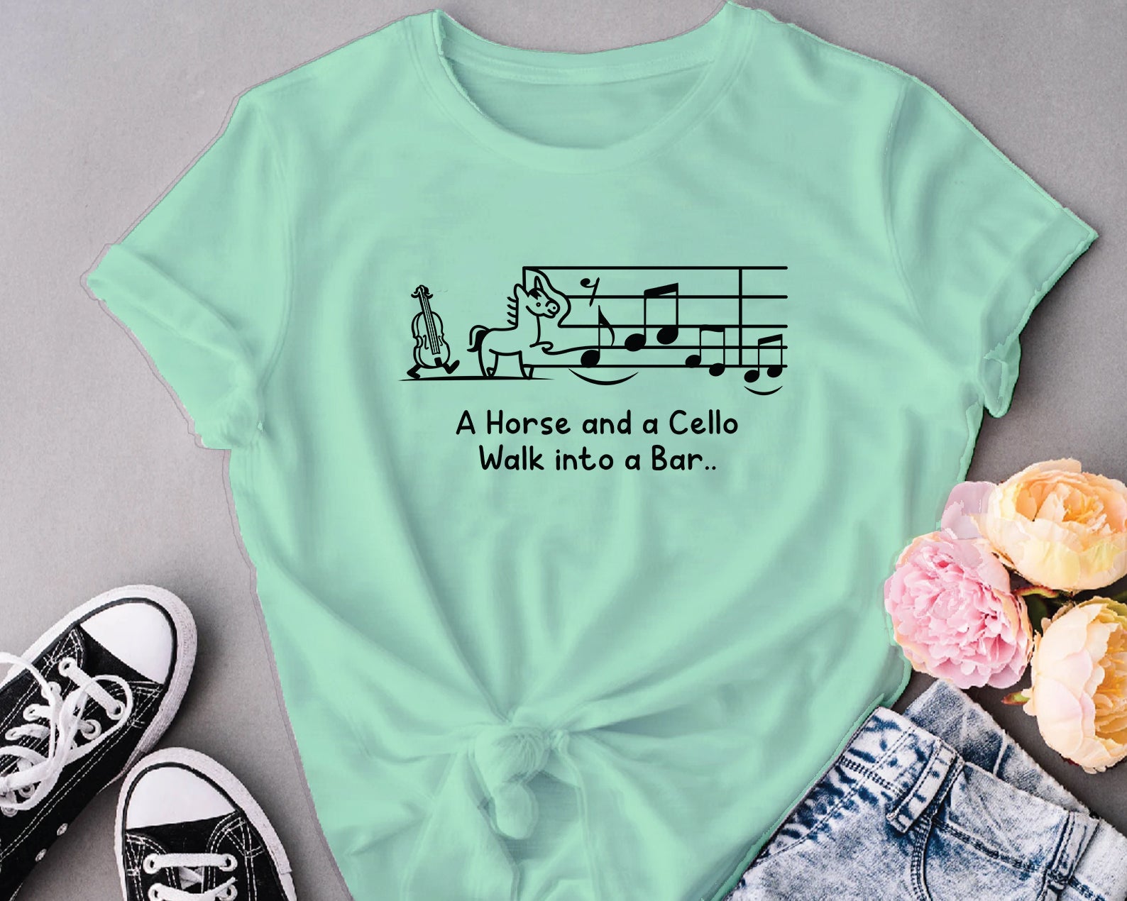 Mint green t-shirt with humorous design and text, placed on a gray surface with flowers and shoes.